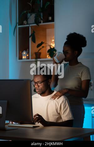 Young african woman supporting husband while he coding on computer and working remotely overtime Stock Photo