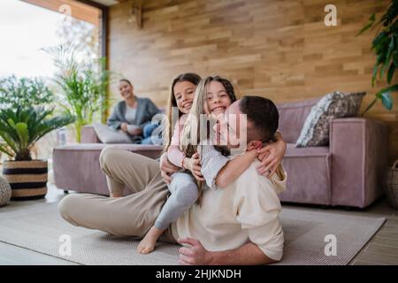 Two happy sisters having fun with father, lying on his back with mother at background at home Stock Photo