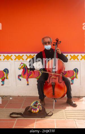 Guatape, Antioquia, Colombia - December 8 2021: Old Brown Man Dressed in Black with Red Suspenders and Sunglasses is Playing the Cello on a Street Stock Photo