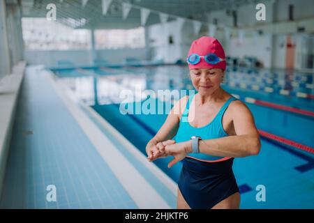 Senior woman setting smartwatch before swim in indoors swimming pool. Stock Photo