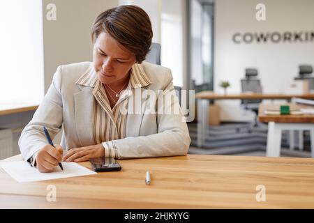 Portrait of adult female manager signing documents while sitting at desk in coworking space, copy space Stock Photo