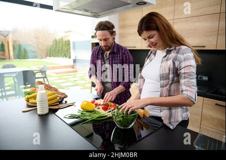 Happy pregnant woman mixing herbs in bowl, standing next to her husband, chopping tomatoes and yellow bell peppers on the kitchen island at home again Stock Photo