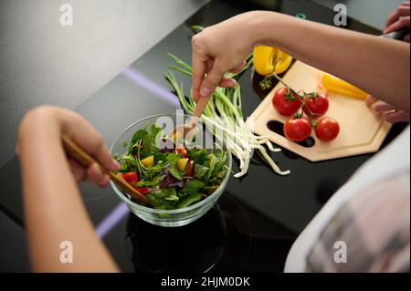 Overhead view of woman's hands holding wooden spoons and mixing ingredients in a glass bowl, preparing delicious healthy salad for dinner in the kitch Stock Photo