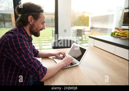 Handsome Caucasian man freelancer in casual attire working remotely on laptop, typing text on laptop with empty blank monitor screen with copy ad spac Stock Photo