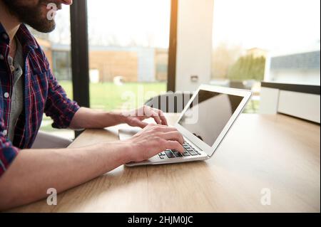 Close-up hands of a man freelancer in casual attire working remotely on laptop, typing text on laptop with empty blank monitor screen with copy ad spa Stock Photo