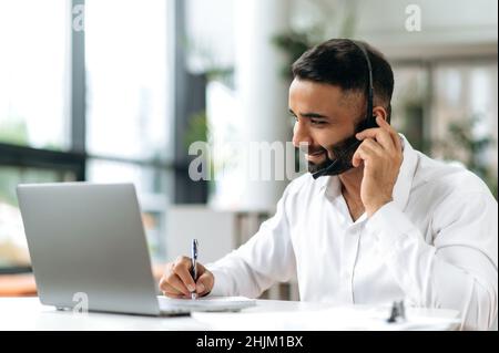 Smart purposeful attractive Indian man with headset, manager, support worker, online consultant, executive sitting at laptop in modern office, listening, recording appeal, doing online consultation Stock Photo