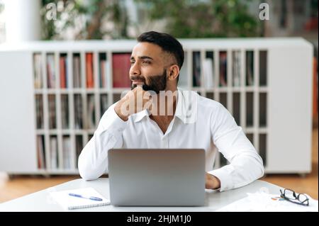Attractive pleasant Indian businessman, mentor, office worker, financial advisor, sitting at the desk in the office, looking away developing a working strategy, analyzing, contemplating business plan Stock Photo