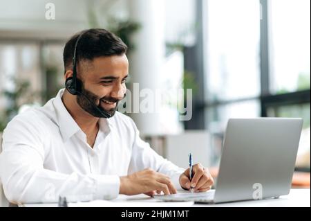 Positive handsome confident Indian man in headphones, manager, support worker, online consultant, director sitting at a laptop in the office, having an online consultation, smiling, takes notes Stock Photo