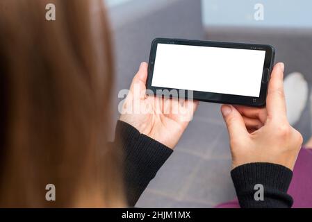 A female is holding and using smartphone with white blank screen. Woman lies on sofa and holds phone with white blank screen. Stock Photo