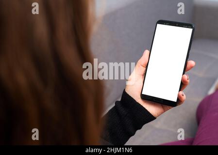 A female is holding and using smartphone with white blank screen. Woman lies on sofa and holds phone with white blank screen. Stock Photo