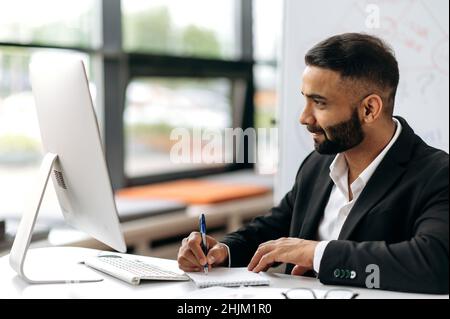 Smart attractive successful Indian businessman in suit, company director, financial advisor or broker sits at his desk, works on laptop, listening online conference, notes information, looks at screen Stock Photo