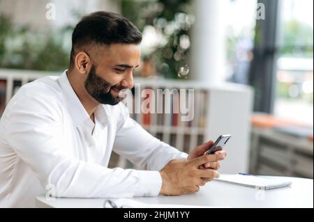 Working texting. A pleasant successful Indian man, a trader, a top manager, sits at his desk, conducts work correspondence on a mobile phone during working hours, smiles friendly Stock Photo