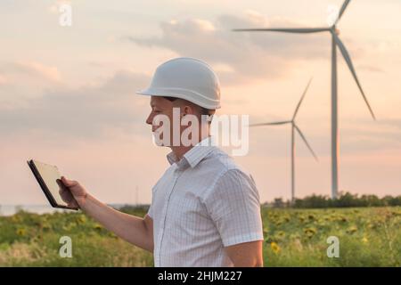 Engineer is checking energy production on wind turbine. Worker in windmills park in helmet and with tablet. Stock Photo