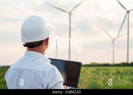 Engineer is checking energy production on wind turbine. Worker in windmills park in helmet and with tablet. Stock Photo