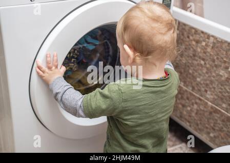 Child boy is looking into the washing machine. Baby boy interested in the cycles of washing machine doing laundry. Stock Photo