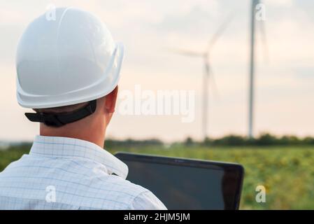 An electrical engineer is using laptop computer, checking on wind turbine energy production. A man with a laptop computer in front of wind turbines. Stock Photo