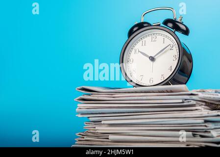 Newspapers and black alarm clock on blue background. Retro alarm clock on newspapers. Media news time concept. Stock Photo