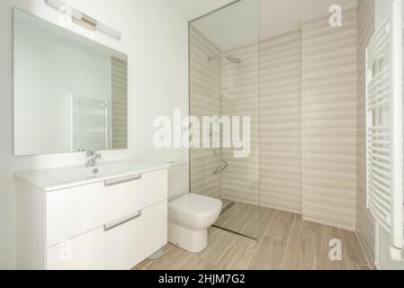 Toilet With white bathroom furniture, sink under the square glass mirror and shower with glass partition with ceramic stoneware floors Stock Photo