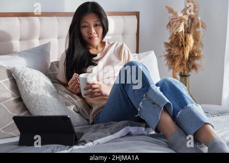 Happy beautiful woman in casual clothes holding cup working on digital tablet sitting on bed Stock Photo