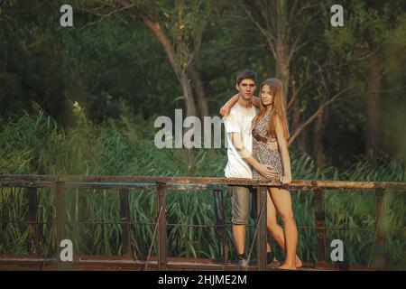 A beautiful couple in nature stands hugging on a wooden bridge over a river overgrown with reeds. Happy lovers celebrate Valentine's Day together. Stock Photo