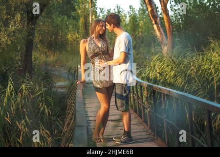 A beautiful couple in nature stands hugging on a wooden bridge over a river overgrown with reeds. Happy lovers celebrate Valentine's Day together. Stock Photo