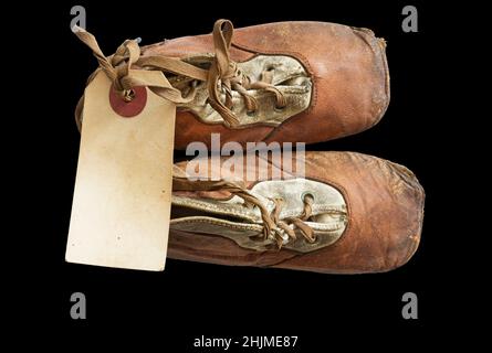 old worn baby shoes from 1908 with a blank paper tag tied onto the laces on black background Stock Photo