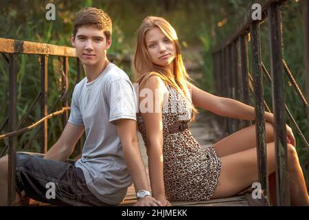 A couple in love sits on a wooden bridge with rusty railings back to back. A happy young couple walks in nature and enjoys communication. Stock Photo
