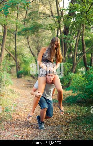 young handsome man with an underage girl are having fun in the forest. Portrait of adult teenagers walking in the park. The girl sits on the shoulders Stock Photo