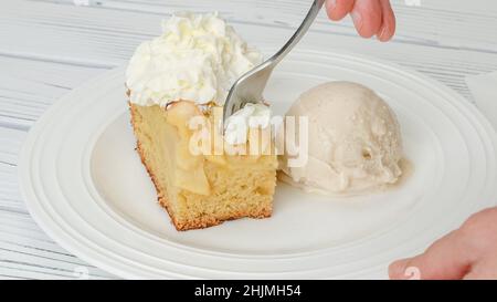 Slice of fresh baked apple cake with biscuit base and sliced apples and whipped cream topping served with vanilla ice cream close up on a plate on whi Stock Photo