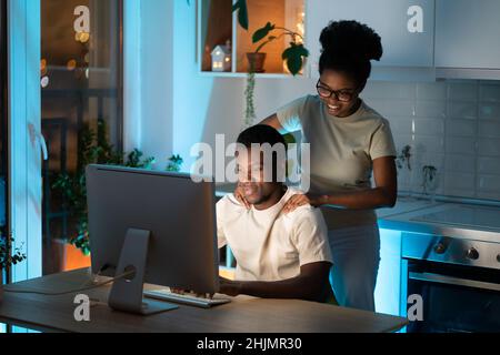 Loving african woman massaging shoulders of husband while he working late on computer at home Stock Photo