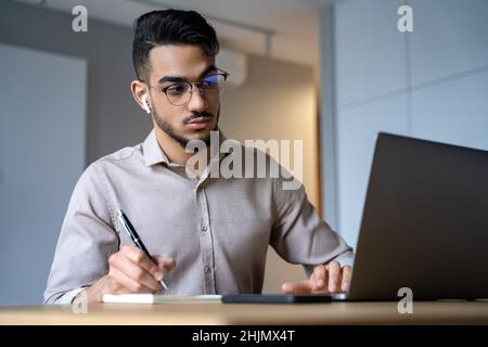 Young indian student at home office watching webinar using laptop making notes Stock Photo