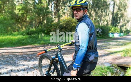 Portrait of mountain biker sitting on his bike wearing helmet, goggles and smartwatch on a country road lined with trees with copy space Stock Photo