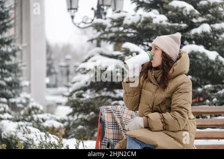 Cheerfully brunette woman drinking from flask outdoors in the city in winter. Warming up, enjoying moment, having brake over snow covered trees Stock Photo