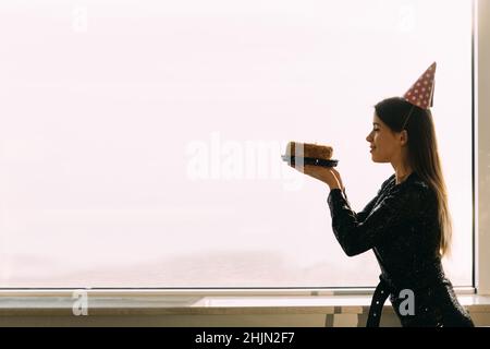 beautiful caucasian woman holding a cake looking at a candle, standing at the office window, light background, profile view of a girl Stock Photo