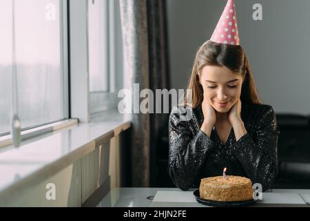 Portrait of a sad young woman sitting at the table on her birthday, a lonely holiday at home, a girl in a cap celebrating an anniversary in quarantine Stock Photo