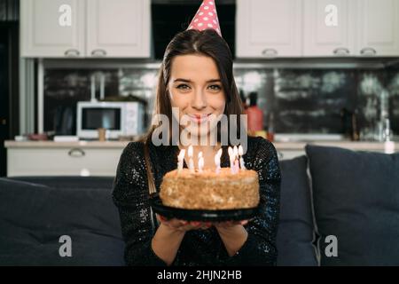 Portrait of a caucasian brunette with a birthday cake with candles, a girl in a cap at home alone celebrates an anniversary Stock Photo