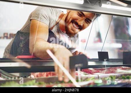 Young man butcher arranging meat products in display case of butcher shop Stock Photo