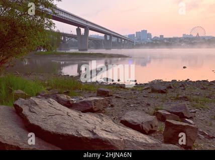 Rocky shore of the Ob in Novosibirsk. Big stones on the riverbank, bridges, a big city with a Ferris wheel on the horizon in the morning fog. Pink sky Stock Photo