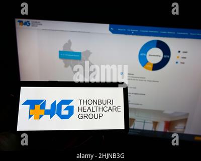 Person holding cellphone with logo of Thai company Thonburi Healthcare Group (THG) on screen in front of business webpage. Focus on phone display. Stock Photo