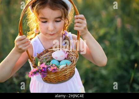 Cute funny girl with painted Easter eggs in spring in nature in a field with golden sunlight and flowers. Easter holiday, Easter bunny with ears, colo Stock Photo