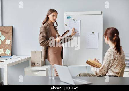 Young pregnant woman pointing at whiteboard presenting her report for colleagues during conference at office Stock Photo