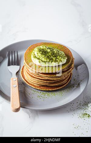 Stack of vegan pancakes with coconut cream and matcha powder on gray plate, white kitchen. Stock Photo