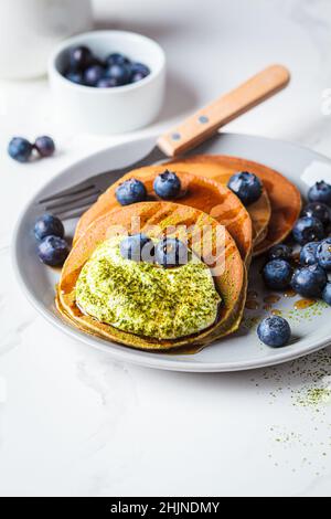 Matcha tea green pancakes with coconut cream, blueberries and maple syrup, white background, copy space. Vegan dessert concept. Stock Photo
