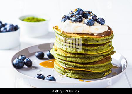 Matcha tea green pancakes with coconut cream, blueberries and maple syrup, white background, copy space. Vegan dessert concept. Stock Photo