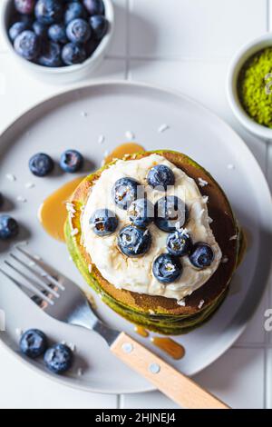 Matcha tea green pancakes with coconut cream, blueberries and maple syrup, white background, top view. Vegan dessert concept. Stock Photo