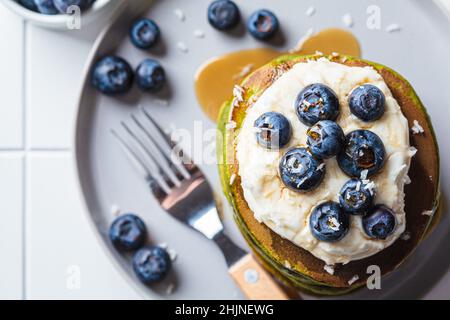 Matcha tea green pancakes with coconut cream, blueberries and maple syrup, white background, top view. Vegan dessert concept. Stock Photo