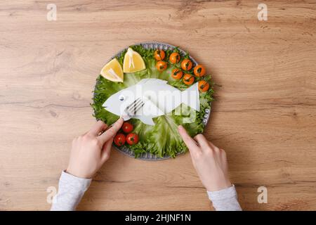 eating plastic fish with vegetables on plate with female hands by fork and knife on wooden background, plastic food, fish pollution concept, earth day Stock Photo