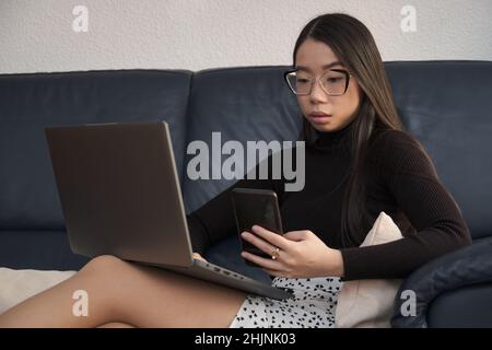Young asian woman using the phone and the laptop sitting on the sofa. Stock Photo
