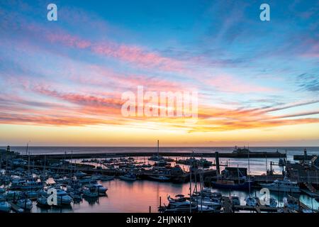 Colourful dawn sky over the English Channel and Ramsgate Royal Harbour in Kent, England. Harbour filled with boats and a yachting marina with an overhead sky of Cirrus fibratus clouds.pink red clouds and yellow sky changing to blue higher up. Stock Photo
