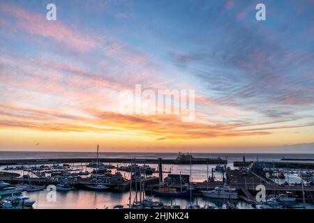 Colourful dawn sky over the English Channel and Ramsgate Royal Harbour in Kent, England. Harbour filled with boats and a yachting marina with an overhead sky of Cirrus fibratus clouds.pink red clouds and yellow sky changing to blue higher up. Stock Photo